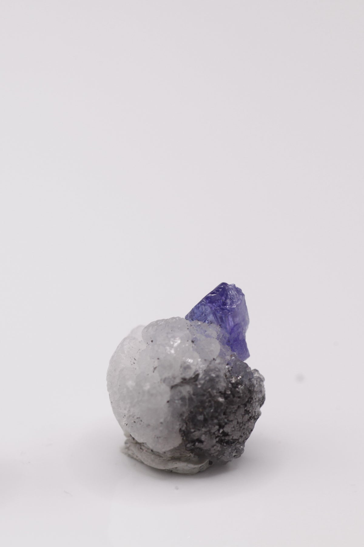 Tanzanite - Mine Mineral Market : A012 Ein kleiner blauer Tansanit-Kristall sitzt auf kugeligem weißen Prehnit-Aggregat. Eine kleine nette Stufe durch den Kontrast zu zusätzlich seitlich anhaftendem Graphit.
