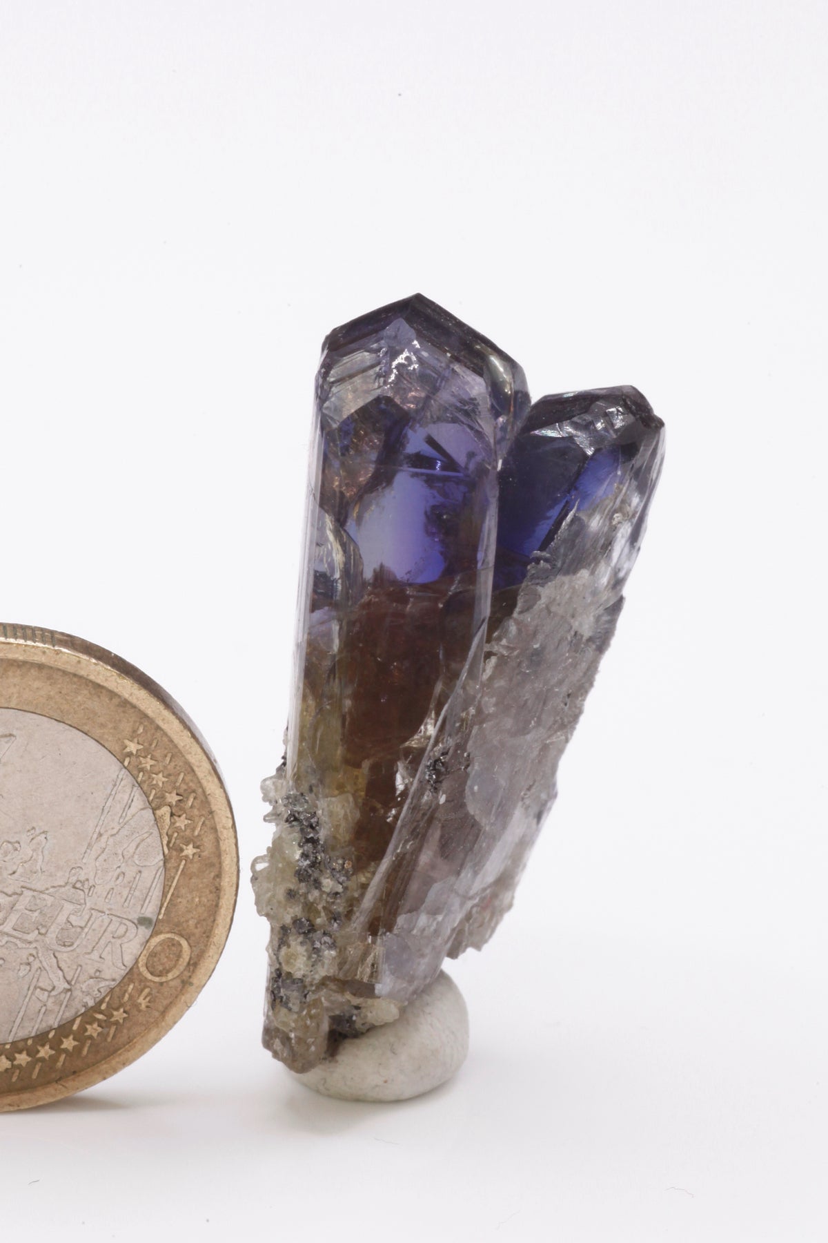 Tanzanite - Mine Mineral Market : A235 Ein schmaler länglicher Doppelkristall, im oberen Bereich mit einem schönen Farbspiel und sehr gutem Glanz. Kleine Partien des Kristalls sind klar. Auf dem unteren Teil etwas Matrix aufsitzend.