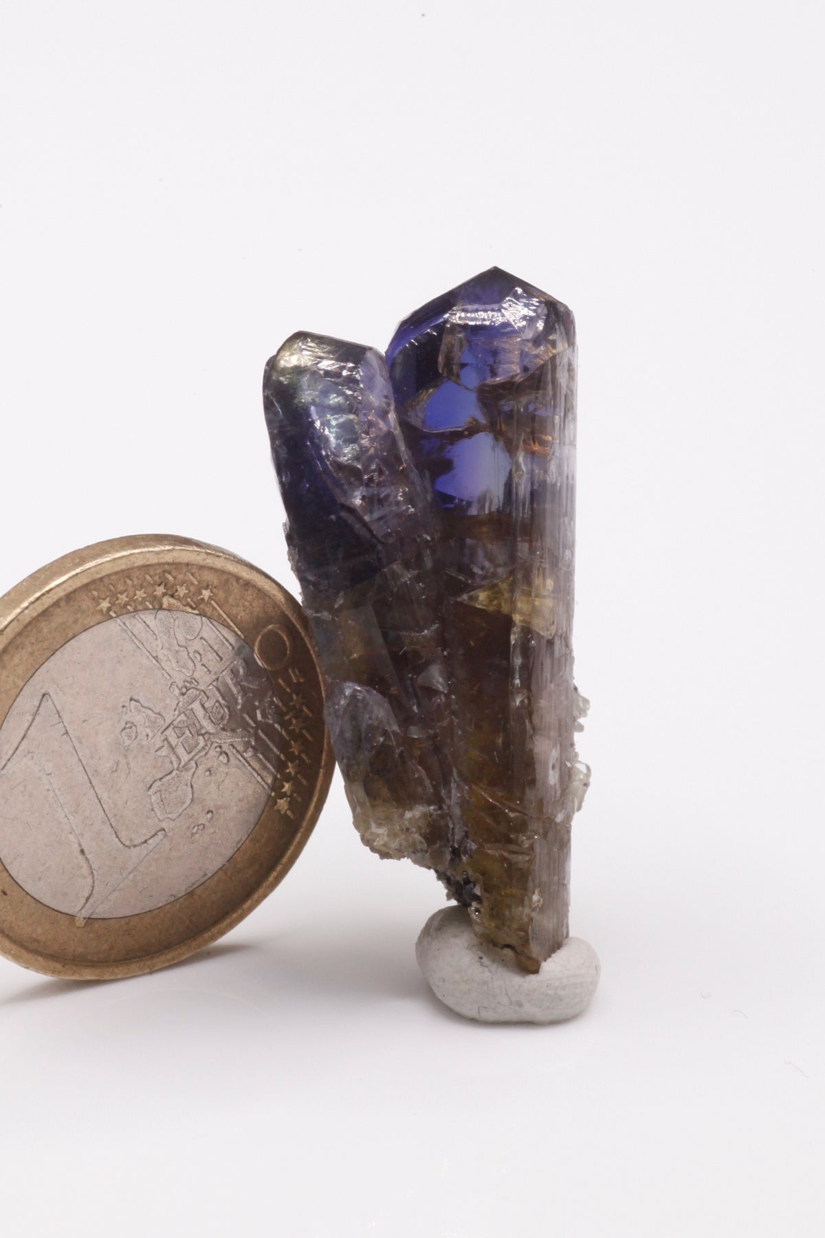 Tanzanite - Mine Mineral Market : A235 Ein schmaler länglicher Doppelkristall, im oberen Bereich mit einem schönen Farbspiel und sehr gutem Glanz. Kleine Partien des Kristalls sind klar. Auf dem unteren Teil etwas Matrix aufsitzend.