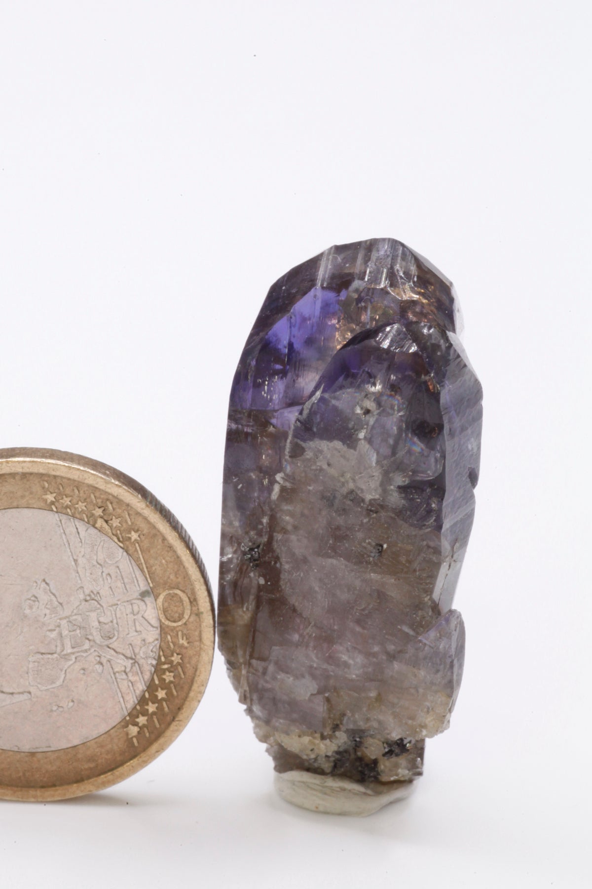 Tanzanite - Mine Mineral Market : A235 Ein schmaler länglicher Doppelkristall, im oberen Bereich mit einem schönen Farbspiel und sehr gutem Glanz. Kleine Partien des Kristalls sind klar. Auf dem unteren Teil etwas Matrix aufsitzend.