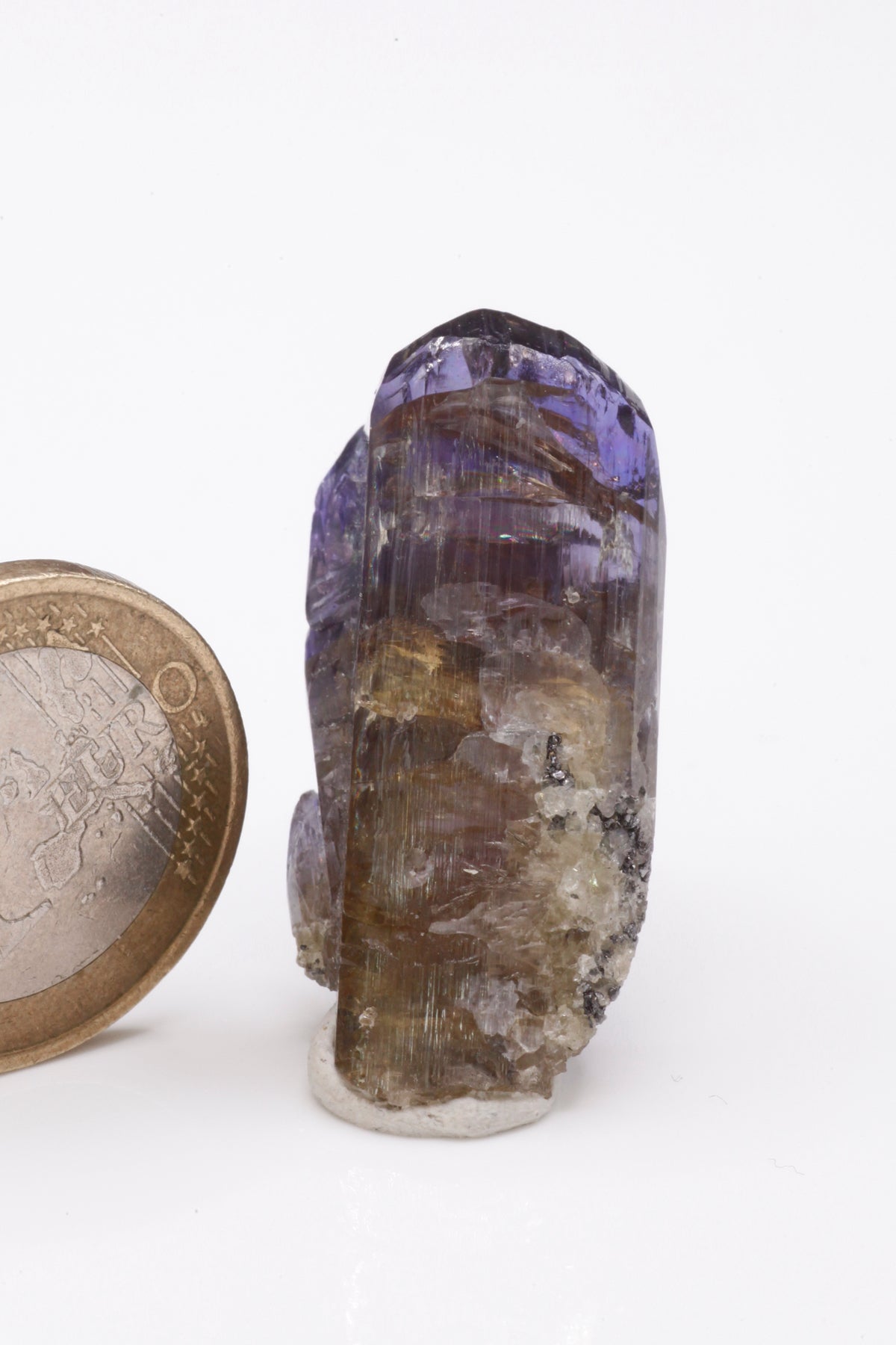 Tanzanite - Mine Mineral Market : A235 Ein schmaler länglicher Doppelkristall, im oberen Bereich mit einem schönen Farbspiel und sehr gutem Glanz. Kleine Partien des Kristalls sind klar. Auf dem unteren Teil etwas Matrix aufsitzend.